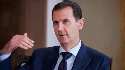 Bashar al-Assad pictured in 2016 speaking while gesturing with one hand. He is wearing a dark suit, white shirt and tie.