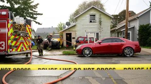 Firefighters work outside a green house with a red car parked in front