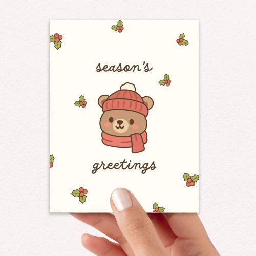 cute Christmas card with a bear wearing a pink hat and scarf, surrounded by holly leaves and berries with text: season's greetings