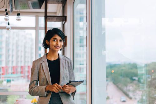 Young Businesswoman Posing by Window Waist up portrait of young female entrepreneur holding digital tablet and smiling happily at camera standing by window professional-woman stock pictures, royalty-free photos & images