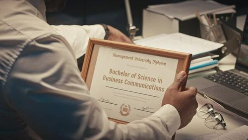 Over shoulder of prosperous African American businessman placing picture frame with bachelor degree diploma in it on office desk at workplace