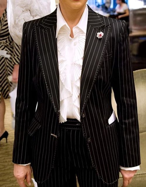 May include: Black pinstripe suit with a white ruffled blouse. The suit features a notched lapel, welt pockets, and a center vent.  The blouse has a pleated ruffle detail at the neckline. A small flower brooch is pinned to the lapel.