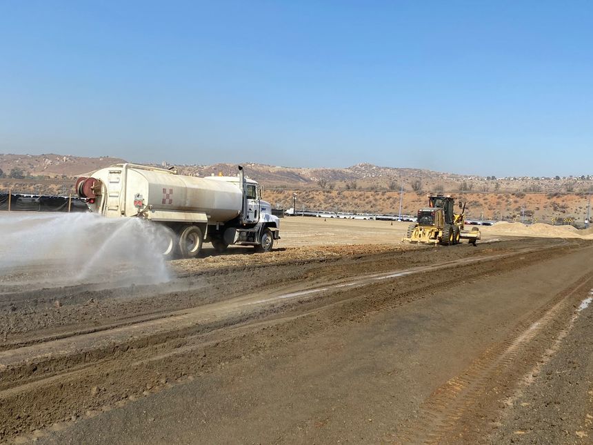 Two construction workers in high-visibility vests and hard hats are operating a large road paver machine. They are laying fresh asphalt on a road, with one worker standing on the machine and the other standing on the ground beside it. The scene is set outdoors with some trees and a wooden fence in the background.