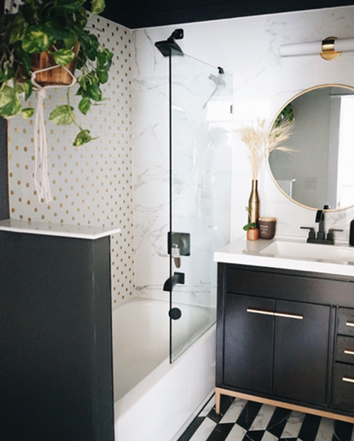 Modern bathroom with a black vanity, round mirror, white countertop, glass shower door, hanging plant, gold polka-dot accent wall, and black-and-white geometric floor tiles.