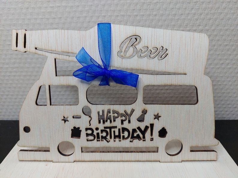 May include: A wooden birthday decoration in the shape of a camper van with a beer bottle on top. The words "Beer" and "Happy Birthday!" are cut out of the wood. A blue ribbon is tied around the beer bottle.