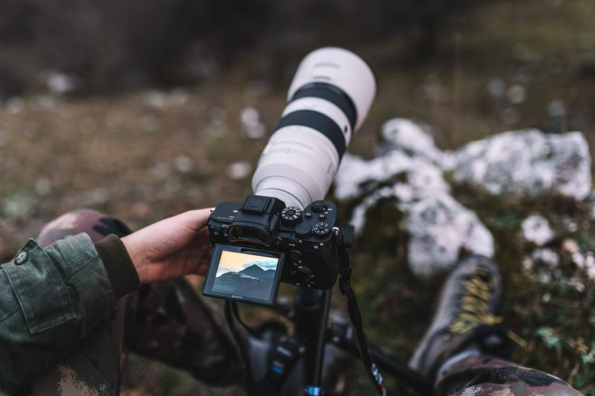 Person holding a dslr camera with a telephoto lens mounted on a tripod outdoors.