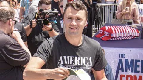 Charlie Kirk smiling at the camera wearig a T shirt with America written accross the front and  crowds iand cameras n the background