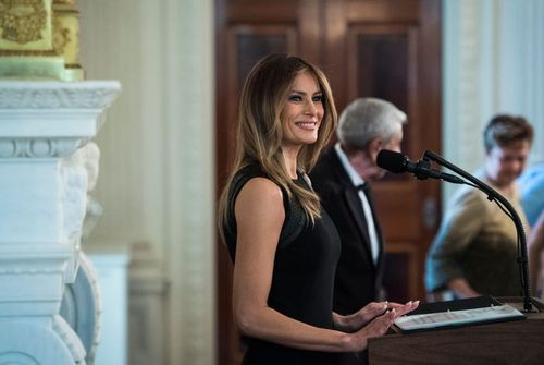 melania trump smiles as she stands at a podium with a microphone attached, she wears a black sleeveless dress