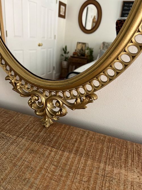 May include: A gold oval mirror with an ornate frame. The frame has a decorative design with a scrollwork pattern and a series of small circles around the edge.