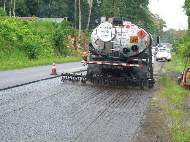 Two construction workers in high-visibility vests and hard hats are operating a large road paver machine. They are laying fresh asphalt on a road, with one worker standing on the machine and the other standing on the ground beside it. The scene is set outdoors with some trees and a wooden fence in the background.