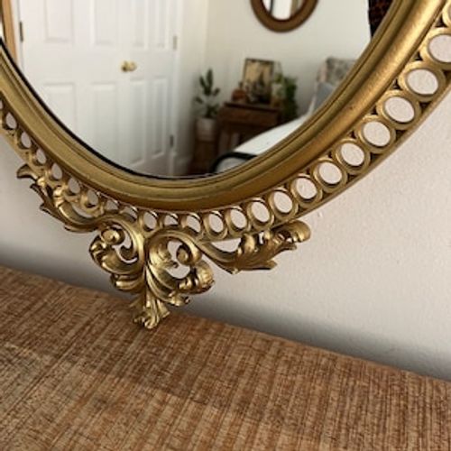 May include: A gold oval mirror with an ornate frame. The frame has a decorative design with a scrollwork pattern and a series of small circles around the edge.