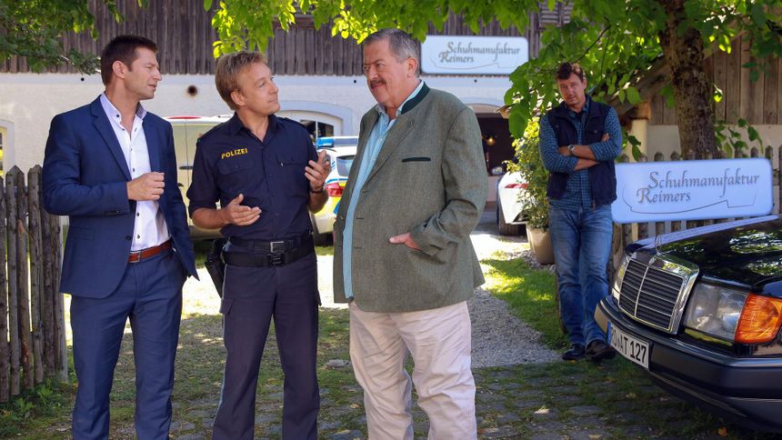 "Die Rosenheim-Cops - Da drückt der Schuh": Kommissar Hansen (Igor Jeftic), Polizist Mohr (Max Müller) und Kommissar Hofer (Joseph Hannesschläger) stehen in der Auffahrt zu einem Haus mit der Aufschrift "Schuhmanufaktur Reimers". Seitlich am Gartenzaun, an dem das gleiche Schild prangt, steht Maik Duschinsky (Markus Langer) mit verschränkten Armen. Er betrachtet die Ermittler.