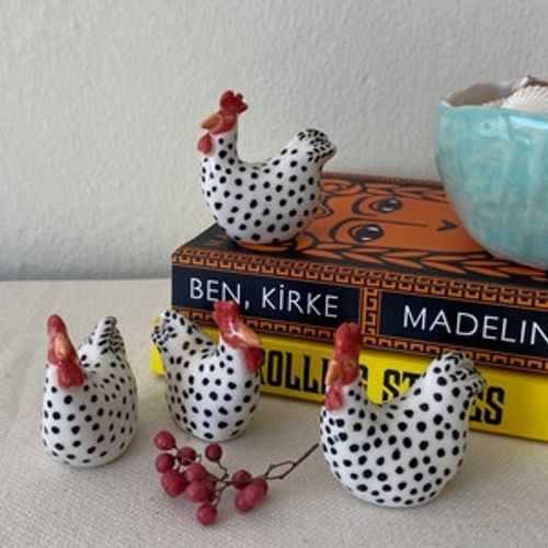 May include: Four black and white ceramic chicken figurines with red combs. The chickens are sitting on a yellow book with the text 'BEN, KIRKE MADELIN' visible. The book is on a white surface.