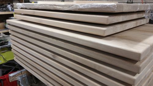 May include: A stack of unfinished wooden tabletops, likely made of oak, with a light brown color. The tabletops are stacked neatly, with the edges aligned.