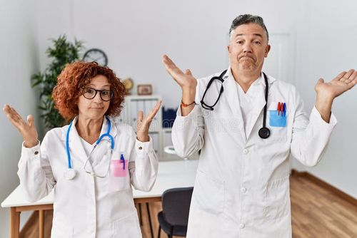 Two middle age doctors at medical clinic clueless and confused expression with arms and hands raised
