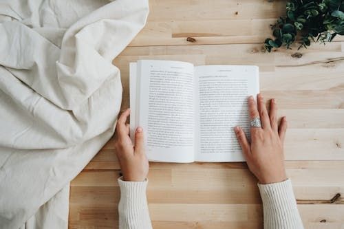 Free Top view of a person reading a book on a wooden table with a cozy setup, ideal for leisure and learning concepts. Stock Photo