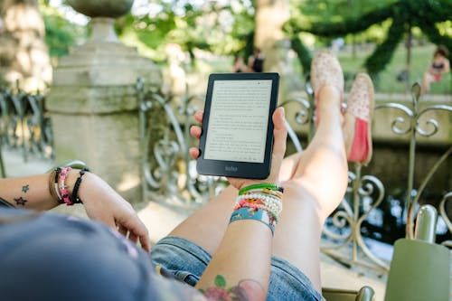 Free A woman leisurely reading a Kobo e-reader outdoors with colorful bracelets and a tattoo visible. Stock Photo