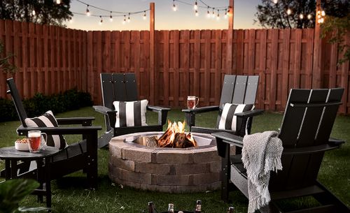 A stone block fire pit with Adirondack chairs in a fenced back yard.