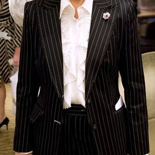 May include: Black pinstripe suit with a white ruffled blouse. The suit features a notched lapel, welt pockets, and a center vent.  The blouse has a pleated ruffle detail at the neckline. A small flower brooch is pinned to the lapel.