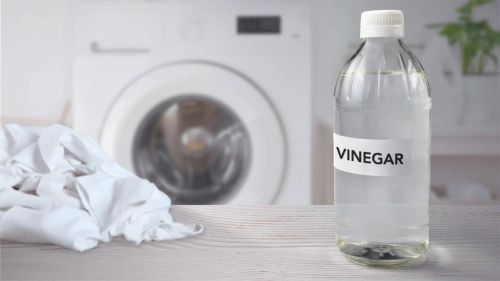 Pile of white laundry and clear glass bottle of white vinegar on wood table, washing machine in background.